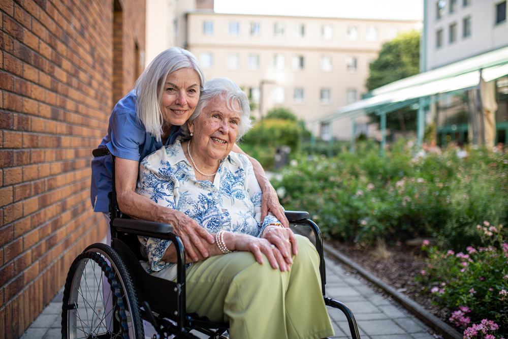 A smiling caregiver hugs an older woman seated in a wheelchair in a sunny garden.